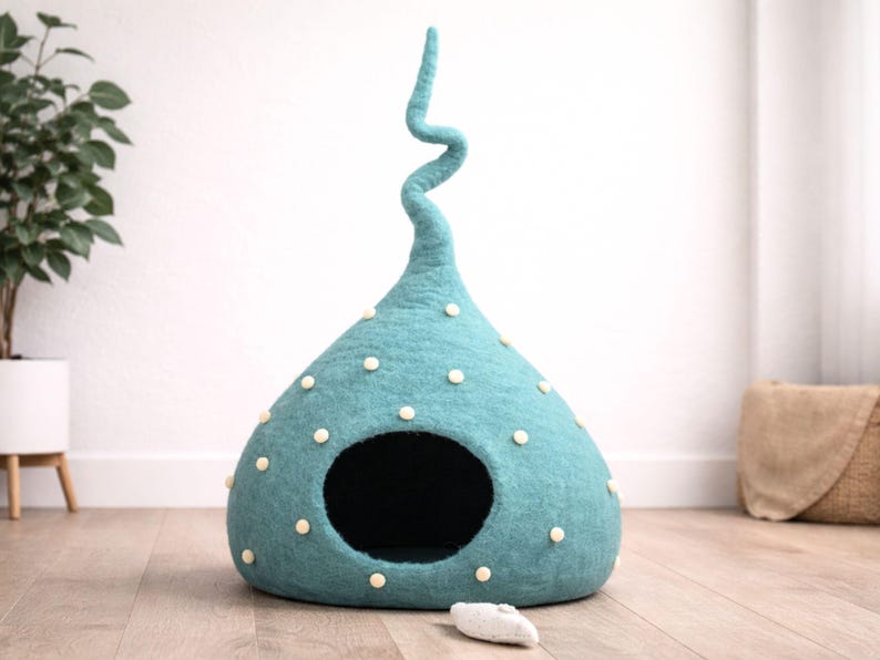 May include: A teal felted pet cave with a tall, spiralling top and a wide, circular opening. The exterior is decorated with small, cream-coloured felted dots scattered across the surface. A small, light-coloured felted toy lies on the floor.