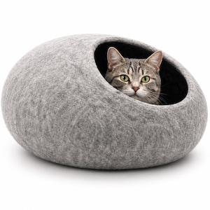 Könnte beinhalten: Eine hellgraue, abgerundete Katzenhöhle aus gefilzter Wolle mit einer glatten, strukturierten Außenseite und einer dunklen Innenöffnung. Eine getigerte Katze mit grünen Augen und grau-braunem Fell sitzt im runden Eingang des Katzenbetts.