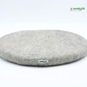 K&ouml;nnte beinhalten: Ein rundes, hellgraues Sitzkissen aus Filz mit einer strukturierten, melierten Oberfl&auml;che. Ein kleines wei&szlig;es Stoffetikett mit dem schwarzen Schriftzug "woolly felt" ist an der Seite angebracht. Das Logo "woolly felt" mit Schaf-Symbol ist sichtbar.