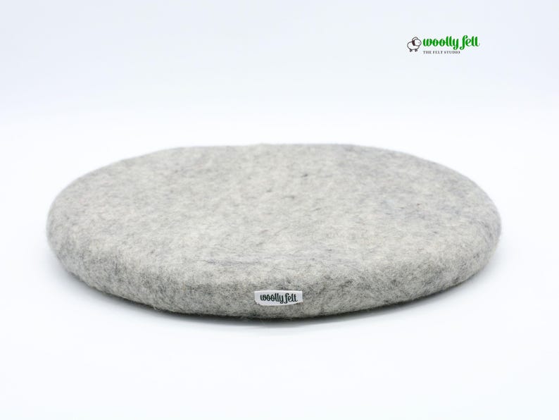 K&ouml;nnte beinhalten: Ein rundes, hellgraues Sitzkissen aus Filz mit einer strukturierten, melierten Oberfl&auml;che. Ein kleines wei&szlig;es Stoffetikett mit dem schwarzen Schriftzug "woolly felt" ist an der Seite angebracht. Das Logo "woolly felt" mit Schaf-Symbol ist sichtbar.