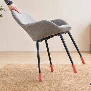 May include: A gray upholstered chair with black metal legs, each fitted with a soft, dusty pink felt floor protector cap. The chair is tilted forward, showcasing the circular felt covers securely attached to the base of all four chair legs.