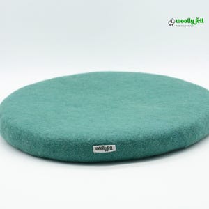 K&ouml;nnte beinhalten: Ein rundes, blaugr&uuml;nes Sitzkissen aus Filz mit einer strukturierten Oberfl&auml;che. Ein kleines wei&szlig;es Stoffetikett mit dem schwarzen Text "woolly felt" ist an der Seite angebracht. Das Markenlogo "woolly felt THE FELT STUDIO" mit einem Schaf-Symbol ist zu sehen.