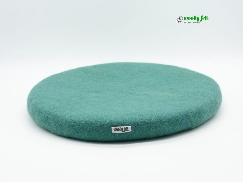 K&ouml;nnte beinhalten: Ein rundes, blaugr&uuml;nes Sitzkissen aus Filz mit einer strukturierten Oberfl&auml;che. Ein kleines wei&szlig;es Stoffetikett mit dem schwarzen Text "woolly felt" ist an der Seite angebracht. Das Markenlogo "woolly felt THE FELT STUDIO" mit einem Schaf-Symbol ist zu sehen.