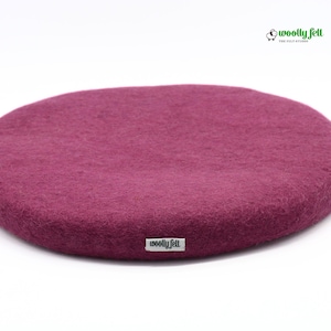 K&ouml;nnte beinhalten: Ein rundes, tief magentafarbenes Sitzkissen aus Filz mit strukturierter Oberfl&auml;che. An der Seite befindet sich ein kleines wei&szlig;es Stoffetikett mit dem schwarzen Schriftzug "woolly felt". Oben rechts ist ein Logo mit Schafsymbol zu sehen.