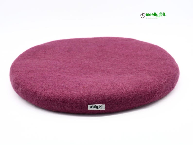 K&ouml;nnte beinhalten: Ein rundes, tief magentafarbenes Sitzkissen aus Filz mit strukturierter Oberfl&auml;che. An der Seite befindet sich ein kleines wei&szlig;es Stoffetikett mit dem schwarzen Schriftzug "woolly felt". Oben rechts ist ein Logo mit Schafsymbol zu sehen.