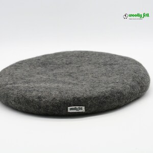 K&ouml;nnte beinhalten: Ein kreisf&ouml;rmiges, dunkelgraues Filzkissen mit einer strukturierten, matten Oberfl&auml;che. Ein kleines wei&szlig;es Stoffetikett mit dem schwarzen Text "woolly felt" ist an der Seite angebracht. Das Logo "woolly felt THE FELT STUDIO" mit Schaf-Symbol ist zu sehen.