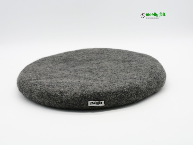 K&ouml;nnte beinhalten: Ein kreisf&ouml;rmiges, dunkelgraues Filzkissen mit einer strukturierten, matten Oberfl&auml;che. Ein kleines wei&szlig;es Stoffetikett mit dem schwarzen Text "woolly felt" ist an der Seite angebracht. Das Logo "woolly felt THE FELT STUDIO" mit Schaf-Symbol ist zu sehen.