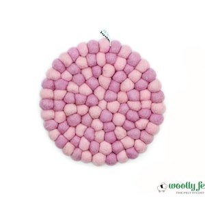 May include: A round trivet made of felted wool balls in shades of pink and lavender. The trivet has a textured, bumpy surface and a small loop for hanging. The Woolly Felt Studio logo is visible in the bottom right corner.
