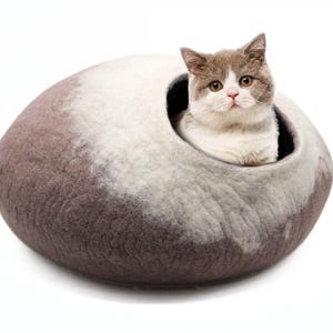 以下が含まれることがあります： 灰色と白のフェルト製の猫用ベッドで、大きな滑らかな石の形をしています。灰色と白の猫がベッドの中に座ってカメラを見ています。