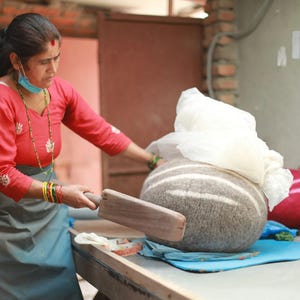 May include: A large, round, grey felted wool object with white horizontal stripes being shaped with a flat wooden paddle. The object is partially covered by a white cloth, and a bright red felted wool item sits nearby on a flat work surface.