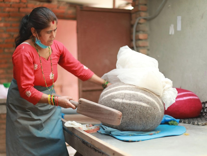 May include: A large, round, grey felted wool object with white horizontal stripes being shaped with a flat wooden paddle. The object is partially covered by a white cloth, and a bright red felted wool item sits nearby on a flat work surface.