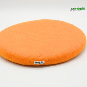 K&ouml;nnte beinhalten: Ein rundes, leuchtend oranges Sitzkissen aus Filz mit einer glatten, dichten Textur. Ein kleines wei&szlig;es Stoffetikett mit dem schwarzen Text "woolly felt" ist an der Seite angebracht. Oben rechts befindet sich ein Logo mit Schafsymbol und Text.