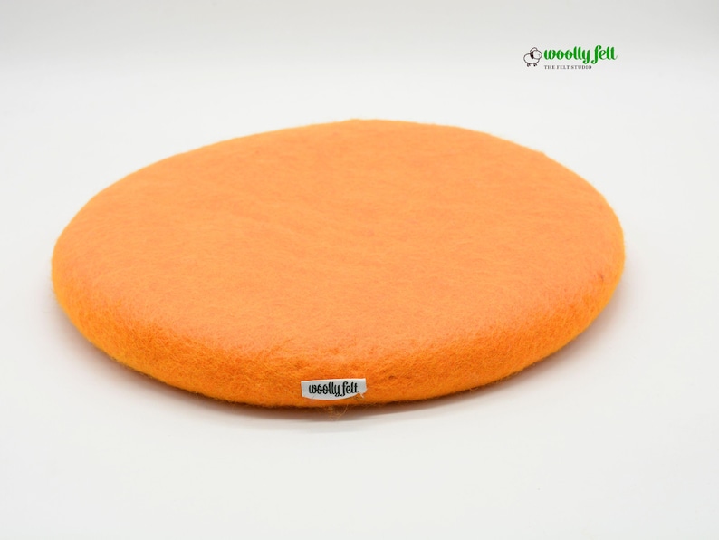 K&ouml;nnte beinhalten: Ein rundes, leuchtend oranges Sitzkissen aus Filz mit einer glatten, dichten Textur. Ein kleines wei&szlig;es Stoffetikett mit dem schwarzen Text "woolly felt" ist an der Seite angebracht. Oben rechts befindet sich ein Logo mit Schafsymbol und Text.