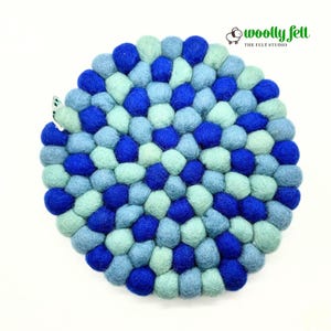 May include: A circular trivet made of numerous small, round felt balls in shades of royal blue, light blue, and mint green. The balls are arranged in a dense, concentric pattern. A small white tag is visible on the left edge. The logo woolly felt THE FELT STUDIO is in the top right corner.