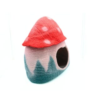 May include: A felt mushroom-shaped pet bed with a red top and white polka dots, a pink base, and a green felt trim. The bed has a round opening for a pet to enter.