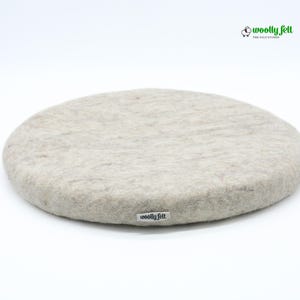K&ouml;nnte beinhalten: Ein rundes Sitzkissen aus beigefarbener Filzwolle mit einer strukturierten, melierten Oberfl&auml;che. An der Seite befindet sich ein kleines wei&szlig;es Stoffetikett mit dem schwarzen Schriftzug "woolly felt". Oben rechts ist ein Logo mit Schaf-Symbol zu sehen.