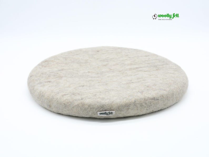 K&ouml;nnte beinhalten: Ein rundes Sitzkissen aus beigefarbener Filzwolle mit einer strukturierten, melierten Oberfl&auml;che. An der Seite befindet sich ein kleines wei&szlig;es Stoffetikett mit dem schwarzen Schriftzug "woolly felt". Oben rechts ist ein Logo mit Schaf-Symbol zu sehen.