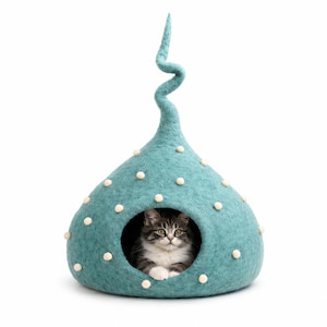 May include: A teal felted cat cave with a tall, spiralling top and small white pom-poms scattered across the exterior. The structure has a circular opening at the front, revealing a fluffy tabby cat with white markings resting inside the enclosure.