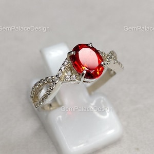 Könnte beinhalten: Ein silberner Ring mit einem großen, oval geschliffenen, roten Edelstein. Das Band hat ein verdrehtes Design, besetzt mit kleinen, klaren Edelsteinen. Der Ring wird auf einem weißen Ständer vor einem weichen, neutralen Hintergrund präsentiert.