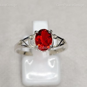Könnte beinhalten: Ein silberner Ring mit einem ovalen, feuerroten Edelstein. Der Ring hat ein verziertes, filigranes Design am Band und um den zentralen Stein. Der Edelstein ist der Blickfang und zeigt eine lebendige, auffällige Farbe. Der Ring wird auf einem weißen Ständer präsentiert.