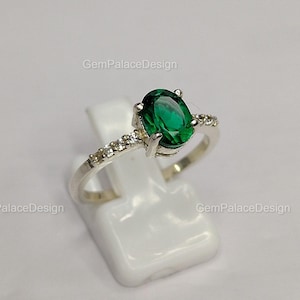 Può includere: Un anello d'argento con una grande pietra verde smeraldo ovale come elemento centrale. La fascia è impreziosita da pietre preziose più piccole e trasparenti. L'anello è esposto su un supporto bianco, che ne mette in risalto il design e la brillantezza.
