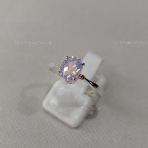 Könnte beinhalten: Ein silberner Ring mit einem ovalen, schillernden Edelstein. Der Edelstein hat sanfte Pastelltöne von Rosa und Lavendel. Der Ring wird auf einem weißen Ständer vor einem neutralen Hintergrund präsentiert.