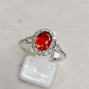 Könnte beinhalten: Ein silberner Ring mit einem leuchtend roten, oval geschliffenen Edelstein, umgeben von einem Kranz aus kleineren, klaren Steinen. Der Ring hat zusätzliche klare Steine am Band, die einen funkelnden Effekt erzeugen. Präsentiert auf einem weißen Ständer.