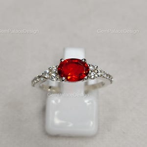 Könnte beinhalten: Ein silberner Ring mit einem großen, oval geschliffenen, feuerroten Edelstein als Mittelpunkt. Das Band ist mit kleineren, klaren Edelsteinen besetzt. Der Ring wird auf einem weißen Ständer präsentiert. Der Text "GemPalaceDesign" ist sichtbar.