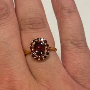 Può includere: Un anello d'oro con una grande pietra ovale rossa circondata da pietre rosse più piccole.