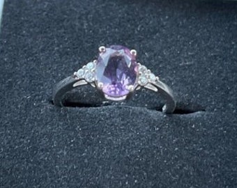 May include: A silver ring with a large oval purple gemstone as the centerpiece, flanked by smaller clear stones. The ring is set in a white box with a black interior, suggesting a jewelry presentation.