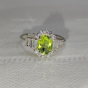 Könnte beinhalten: Ein silberfarbener Ring mit einem zentralen, oval geschliffenen, lindgrünen Peridot-Edelstein. Der Stein ist von einem Kranz aus kleinen runden weißen Steinen umgeben, wobei auf jeder Seite der Ringschiene drei weiße Baguette-Steine sitzen.
