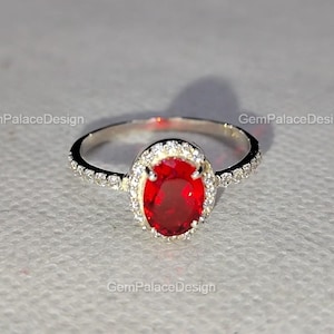 Könnte beinhalten: Ein silberner Ring mit einem großen, oval geschliffenen roten Edelstein in der Mitte, umgeben von einem Halo aus kleineren, klaren Edelsteinen. Der Ring ist vor einem neutralen Hintergrund abgebildet.