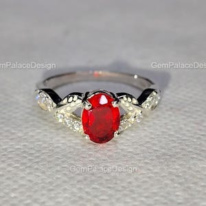 Könnte beinhalten: Ein silberner Ring mit einem ovalen, tiefroten Edelstein im Zentrum. Das Band hat ein verdrehtes Design mit kleineren, klaren Edelsteinen an den Seiten. Der Text "GemPalaceDesign" ist wiederholt im Bild zu sehen.