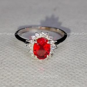Könnte beinhalten: Ein silberner Ring mit einem großen, oval geschliffenen, roten Edelstein, umgeben von kleineren, klaren Steinen. Der Ring hat ein schmales Band. Im Hintergrund ist der Text "GemPalaceDesign" zu sehen.