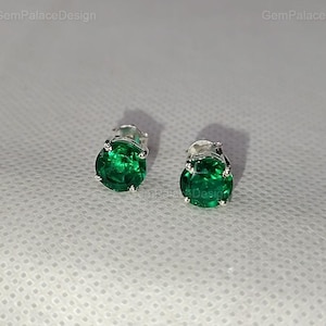 Könnte beinhalten: Ein Paar Ohrstecker mit leuchtend grünen Edelsteinen, die in silberfarbenem Metall gefasst sind. Die Edelsteine haben einen runden Schliff und werden vor einem weichen, strukturierten Hintergrund präsentiert. Klassischer Schmuck.