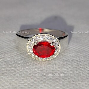Könnte beinhalten: Ein silberner Ring mit einem großen, ovalen, tiefroten Edelstein, umgeben von einem Halo aus kleineren, klaren Steinen. Der Ring hat einen breiten, glatten Band. Das Bild ist gut beleuchtet und hebt die Details und das Funkeln des Rings hervor. Es ist ein Schmuckstück.