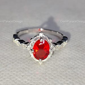 Könnte beinhalten: Ein silberner Ring mit einem ovalen, leuchtend roten Edelstein in der Mitte. Der Ring hat eine dekorative Fassung mit kleinen schwarzen und klaren Akzenten. Der Ring ist vor einem weißen Hintergrund platziert.