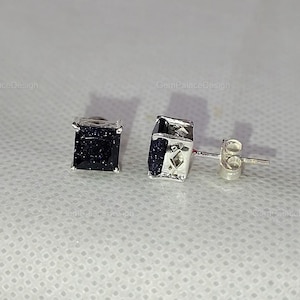 Könnte beinhalten: Ein Paar quadratische Ohrstecker mit einem dunkelblauen, glitzernden Stein, der in einen silbernen Metallrahmen gefasst ist. Die Ohrringe werden auf einer weißen, strukturierten Oberfläche präsentiert.