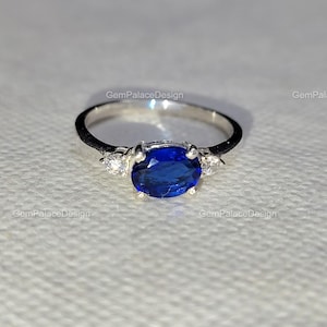 Könnte beinhalten: Ein silberner Ring mit einem großen, oval geschliffenen, tiefblauen Edelstein als Mittelpunkt. Zwei kleinere, klare Edelsteine flankieren den Hauptstein. Der Ring liegt auf einer weißen Oberfläche.