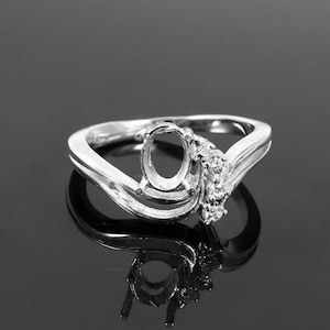 May include: A silver ring with an oval-shaped setting and a delicate design of small stones. The ring has a curved band and a wave-like design.