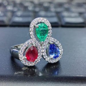 Könnte beinhalten: Ein silberner Ring mit drei birnenförmigen Edelsteinen: einem grünen Smaragd, einem roten Rubin und einem blauen Saphir. Die Edelsteine sind in einer Weißgoldfassung mit einem Diamantenhalo um jeden Stein gefasst.