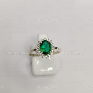 Könnte beinhalten: Ein silberner Ring mit einem ovalen, leuchtend grünen Edelstein, umgeben von einem Kranz aus kleineren, klaren Edelsteinen. Der Ring wird auf einem weißen Ständer präsentiert, der sein elegantes Design hervorhebt. Dies ist ein Schmuckstück.