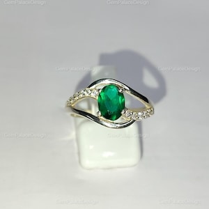 Pode incluir: Um anel de prata com uma pedra preciosa verde esmeralda oval no centro. A banda tem um design único com pequenas pedras claras nas laterais. O anel é exibido em um suporte branco.