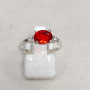 Könnte beinhalten: Ein silberner Ring mit einem großen, oval geschliffenen, feuerroten Edelstein als Mittelpunkt. Zwei kleinere, klare Edelsteine flankieren den Hauptstein. Der Ring wird auf einem weißen Ständer präsentiert, um sein Design hervorzuheben.