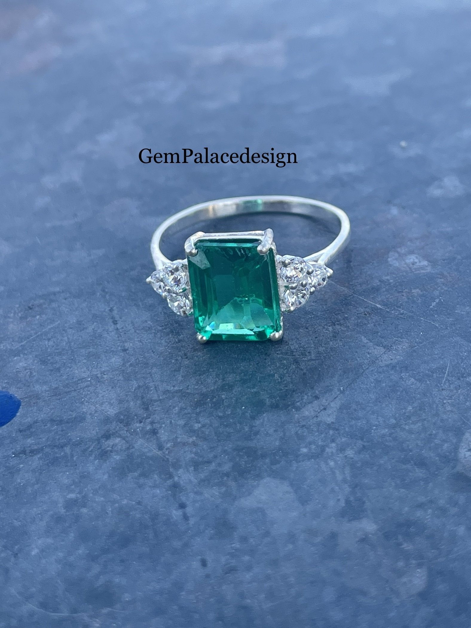 May include: A silver ring featuring a large, rectangular emerald-cut green gemstone. The ring has smaller, clear gemstones on either side of the main stone. The text "GemPalacedesign" is visible in the image.