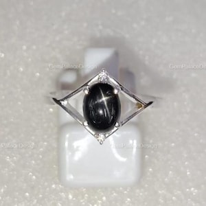 Peut inclure: Bague en argent ornée d'une pierre précieuse saphir étoilé noir. La pierre ovale est sertie dans un cadre géométrique avec de petits diamants. La bague est présentée sur une surface blanche.