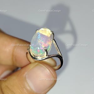 Könnte beinhalten: Ein Silberring mit einem großen, ovalen Opal-Edelstein. Der Opal zeigt ein lebendiges Farbspiel mit grünen, blauen und roten Blitzen. Der Ring hat ein schlichtes, poliertes Silberdesign.