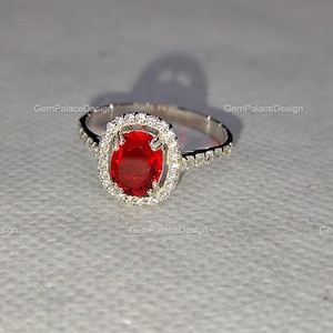 Könnte beinhalten: Ein silberner Ring mit einem großen, oval geschliffenen, tiefroten Edelstein, umgeben von einem Halo aus kleineren, klaren Steinen. Das Ringband ist ebenfalls mit kleinen, klaren Steinen verziert. Der Text "GemPalaceDesign" ist auf dem Bild aufgedruckt.