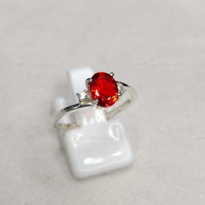 Könnte beinhalten: Ein silberner Ring mit einem großen, oval geschliffenen, leuchtend roten Edelstein als Mittelpunkt. Zwei kleinere, klare Edelsteine flankieren den Hauptstein. Der Ring wird auf einem weißen Ständer vor neutralem Hintergrund präsentiert.