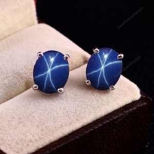 Pode incluir: Um par de brincos de prata com pedras preciosas de safira azul estrela. As pedras preciosas são de forma oval e têm um padrão de estrela.